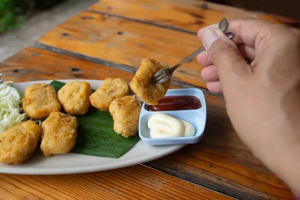 Someone hand uses fork to get a picec of chicken nuggets from disk on wooden table.