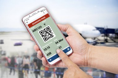 Birisi elinde akıllı telefonuyla uçak biletini ve arka planda bekleyen bir sürü insanı gösteriyor..