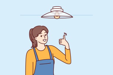 Kadın elektrikçi avizeyi tamir ettikten ya da tavan altında ampulü değiştirdikten sonra başparmağını kaldırmış. Tulumlu kız elektrikçi olarak çalışıyor. Elektriği kurma görevlerini başarıyla yerine getiriyor.