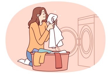 Ev kadını çamaşır makinesinin yanında oturur ve temiz çamaşır deterjanı kullandıktan sonra temiz kokulu havluyu içine çeker. Çamaşır makinesinden çamaşırları çıkarıp kaseye koyuyor.