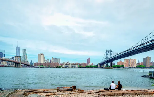 ニューヨーク アメリカ 2014年7月16日 若いカップルが川沿いに座り 下のマンハッタンのスカイラインを楽しんでいます ブルックリン橋は左側 右側はマンハッタン橋 ストック画像