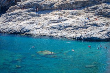 Yunanistan 'ın Amorgos kentindeki Mouros plajı - 25 Temmuz 2024: Yüzücüler denizde ve plajın uzak ucundaki kayalıklarda. Mouros plajı Amorgos, Cyclades, Yunanistan 'da inanılmaz bir çakıl taşı plajı..