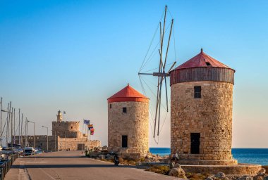 Rodos, Yunanistan - 20 Ağustos 2016: Mandraki limanındaki uzun dalga kırıcıda ortaçağ rüzgar değirmenleri. Arka planda Aziz Niklas kalesi ve deniz feneri var..