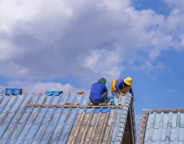 Sızdıran çıkıntıları onaran işçiler çatıyı tamir ediyor ve mavi gökyüzü arka planındaki kırık kiremit çatısını değiştiriyor.
