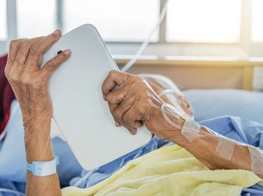 Hastanenin koğuşunda elinde serumla yatarken elinde dijital tablet olan yaşlı hastayı kapat. 
