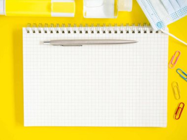 Üst manzara, düz yatış, ofis sarısı masa üstünde boş defter kalem, el dezenfektanı alkol jeli, tıbbi maske. Metnin için alanı kopyala. İş ve eğitim kavramı.