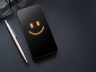 Deneyimler, geri bildirim, değerlendirme ve memnuniyet anketi konsepti. Siyah çalışma masasının üzerinde gülen bir simgeyle cep telefonu ekranı, kalem, gözlük..