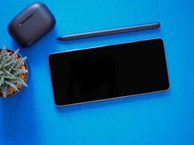 Üst görüş, boş bir akıllı telefon ekranı ve kablosuz kulaklık, mavi arka planda bir kalem. Şablon, ürünün metin ve logosu için cep telefonu boş ekran maketi.