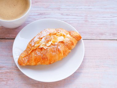 Croissant with cream and almonds on white circle plate and coffee cup over wooden table. Close up, space for text