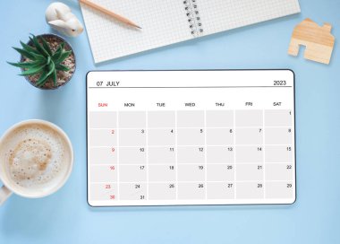 Üst manzara, Temmuz 2023 takvim uygulaması sayfasını gösteren tablet bilgisayar ve kaktüs, kahve fincanı, kalem, mavi arka planda bir tavşan heykeli. Zamanlama planlaması için takvim uygulaması