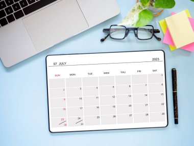 Üst görünüm, düz tablet bilgisayar Temmuz 2023 takvim uygulama sayfası ve dizüstü bilgisayar, kalem ve gözlükleri mavi arkaplanda gösteriyor. Zamanlama planlaması için takvim uygulaması