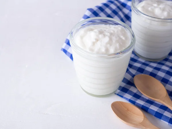 Greek yogurt in cup glass and checkered blue cloth, wooden spoon on white wooden background.
