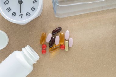 Üst manzara, dağınık destek hapları ve kapsüller ve beyaz plastik hap şişesi, saat, kahverengi arka planda şişe su. Sağlık, sağlık ve bakım farmakolojisi kavramı