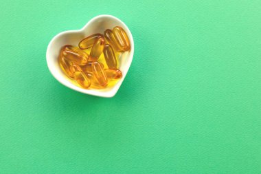 Üst manzara, beyaz, kalp şeklinde bir kase dolusu saydam sarı kapsül ilacı ya da parlak yeşil arka plana karşı takviyeler..