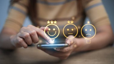Customer service evaluation concept. smiling Asian female Is using a smartphone And she is pressing face emoticon smile in satisfaction on virtual touch screen.