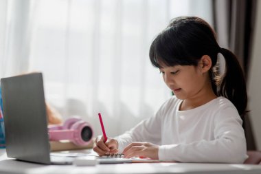 Asyalı liseli kız evde dizüstü bilgisayarla ödevini yapıyor. Çocuklar çalışmak için aletler kullanırlar. Çocuklar için eğitim ve uzak mesafe eğitimi. Karantina sırasında evde eğitim. Evde kal.