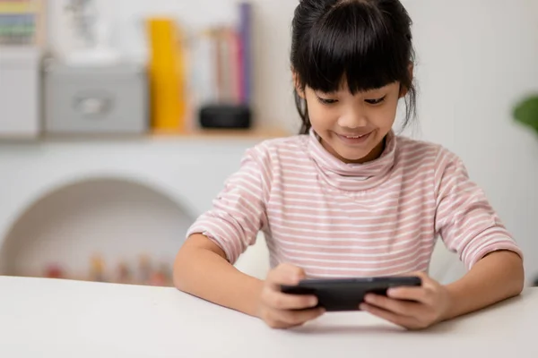 Cute little girl uses smartphone while sitting at the sofa in the living room. Child surfing the internet on mobile phone, browses through internet and watches cartoons online at home