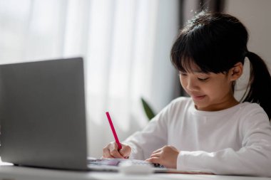 Asyalı liseli kız evde dizüstü bilgisayarla ödevini yapıyor. Çocuklar çalışmak için aletler kullanırlar. Çocuklar için eğitim ve uzak mesafe eğitimi. Karantina sırasında evde eğitim. Evde kal.