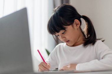 Asyalı liseli kız evde dizüstü bilgisayarla ödevini yapıyor. Çocuklar çalışmak için aletler kullanırlar. Çocuklar için eğitim ve uzak mesafe eğitimi. Karantina sırasında evde eğitim. Evde kal.