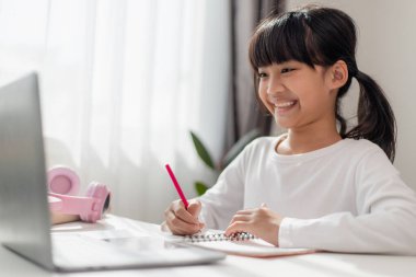 Asyalı liseli kız evde dizüstü bilgisayarla ödevini yapıyor. Çocuklar çalışmak için aletler kullanırlar. Çocuklar için eğitim ve uzak mesafe eğitimi. Karantina sırasında evde eğitim. Evde kal.