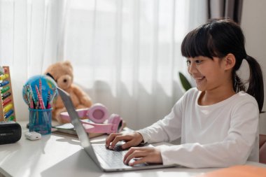 Asyalı liseli kız evde dizüstü bilgisayarla ödevini yapıyor. Çocuklar çalışmak için aletler kullanırlar. Çocuklar için eğitim ve uzak mesafe eğitimi. Karantina sırasında evde eğitim. Evde kal.