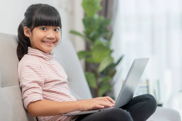 Asian Little school kid girl use laptop computer sitting on sofa alone at home. Child learning reading online social media content, play education lessons game chatting with friends. Children tech addiction