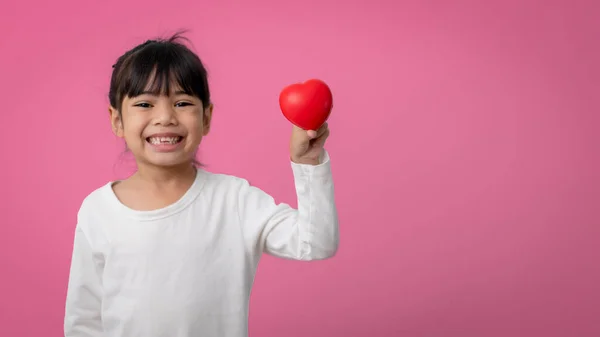 Little Asian girl holding red heart shaped application and looking at camera happily on pink.