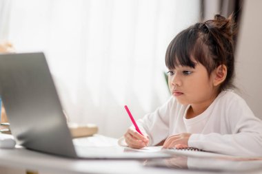 Asyalı liseli kız evde dizüstü bilgisayarla ödevini yapıyor. Çocuklar çalışmak için aletler kullanırlar. Çocuklar için eğitim ve uzak mesafe eğitimi. Karantina sırasında evde eğitim. Evde kal.
