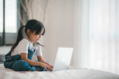 Asyalı kız çocuk yatakta oturuyor ve internette internetten öğreniyor. Evde yalnızlık çekerken videoyla ilgili sanal sınıf dersi. Uzaktan kumandalı video eğitimi. Çocuklar için modern okul eğitimi