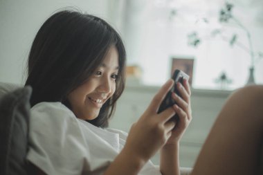 Mutlu Asyalı kız evde rahat bir kanepede rahat bir şekilde mesajlaşıyor ve mesajlaşıyor, gülümseyen kız cep telefonu kullanıyor, internette mesajlaşıyor ve evden online alışveriş yapıyor..