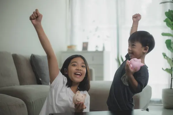 Little Asian girl and boy saving money in a piggy bank, learning about saving, The Kid saves money for future education. Money, finances, insurance, and people concepts.