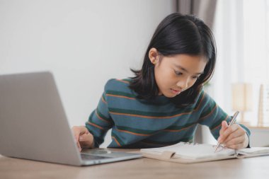 Bir kız dizüstü bilgisayarla masada otururken bir deftere yazıyor. O, işine odaklanmış ve ciddi bir ruh hali içinde.