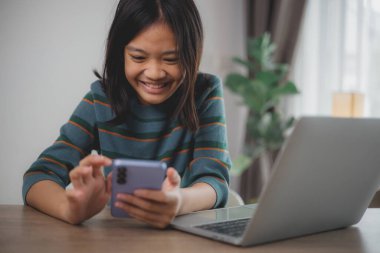Genç bir kız dizüstü bilgisayarı ve cep telefonu olan bir masada oturuyor. Cep telefonuna bakıp gülümsüyor.