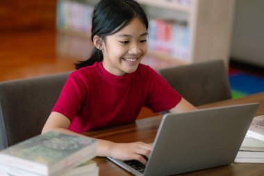 Genç bir kız bir dizüstü bilgisayar ve birkaç kitapla bir masada oturuyor. Gülümsüyor ve eğleniyor.