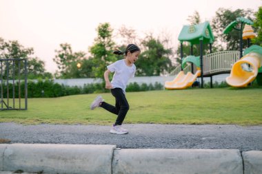 Genç bir kız oyun parkının yanındaki kaldırımda koşuyor. Beyaz gömlek ve siyah pantolon giyiyor.