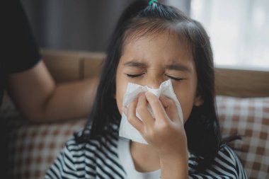 Bir kız burnunu çekiyor ve burnunu mendille siliyor. Kanepede oturuyor.