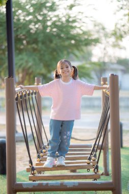 Genç bir kız ahşap bir köprüden geçiyor. Pembe bir gömlek ve kot pantolon giyiyor.