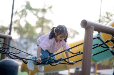 Küçük bir kız çocuk parkında oynuyor. O bir ip merdivene tırmanıyor. Oyun alanı diğer çocuklarla dolu.