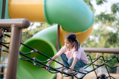 Küçük bir kız çocuk parkında oynuyor. O bir ip merdivene tırmanıyor. Oyun alanı diğer çocuklarla dolu.