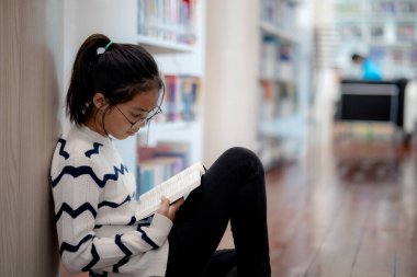 Bir kız yerde oturuyor ve kitap okuyor. Gözlük ve çizgili bir kazak giyiyor.
