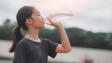 Şişeden su içen bir kız. Kız siyah bir gömlek giyiyor ve saçları at kuyruğu.