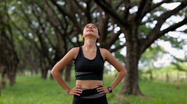 Bir kadın ormanda dikiliyor, gökyüzüne bakıyor. Siyah bir atlet ve siyah tayt giyiyor.