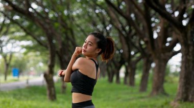 Bir kadın bir direğin yanındaki kaldırımda bacağını esnetiyor. Direk yola yakın ve arka planda ağaçlar var.