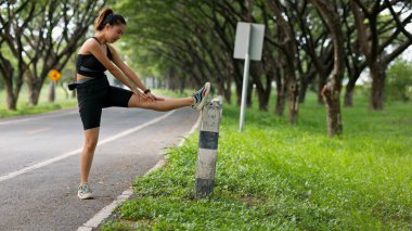 Bir kadın yolun kenarında bacağını esnetiyor. Yol ağaçlarla kaplı ve çimenlerin üzerinde bir direk var.