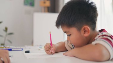 Bir çocuk bir kağıt parçasına pembe bir kalemle yazıyor. O işine odaklanmış ve eğleniyor gibi görünüyor. Sahne penceresi olan bir odada geçiyor.