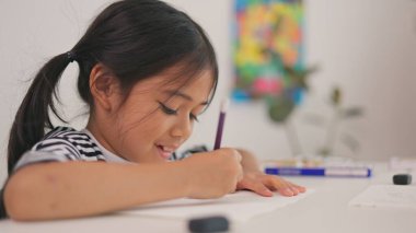 Genç bir kız bir kağıt parçasına mor bir kalemle yazıyor. Gülümsüyor ve eğleniyor. Sahne pozitif ve mutlu bir atmosferi gösteriyor.