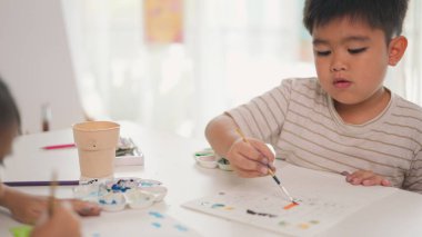 Bir çocuk yanında bir bardak suyla bir resim çiziyor. Çizgili bir gömlek giyiyor.