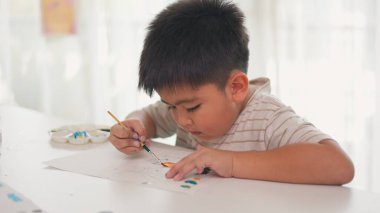 Genç bir çocuk beyaz bir masanın üzerine resim çiziyor. O bir fırça ve renk paleti kullanıyor. Çocuk sanatına odaklanmış gibi görünüyor, ve sahne yaratıcılık ve keyif hissini aktarıyor.