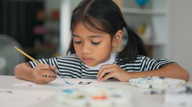 Genç bir kız fırçayla bir resim yapıyor. Çizgili bir gömlek giyiyor ve saçları örgülü saçı var. Resim beyaz bir yüzeyde ve o renkli ve yaratıcı bir sanat eseri.