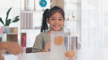 Genç bir kız bir oğlanın önünde bir oyuncakla oynuyor. Çocuk elinde bir kupa ve kâseyle bir masada oturuyor.